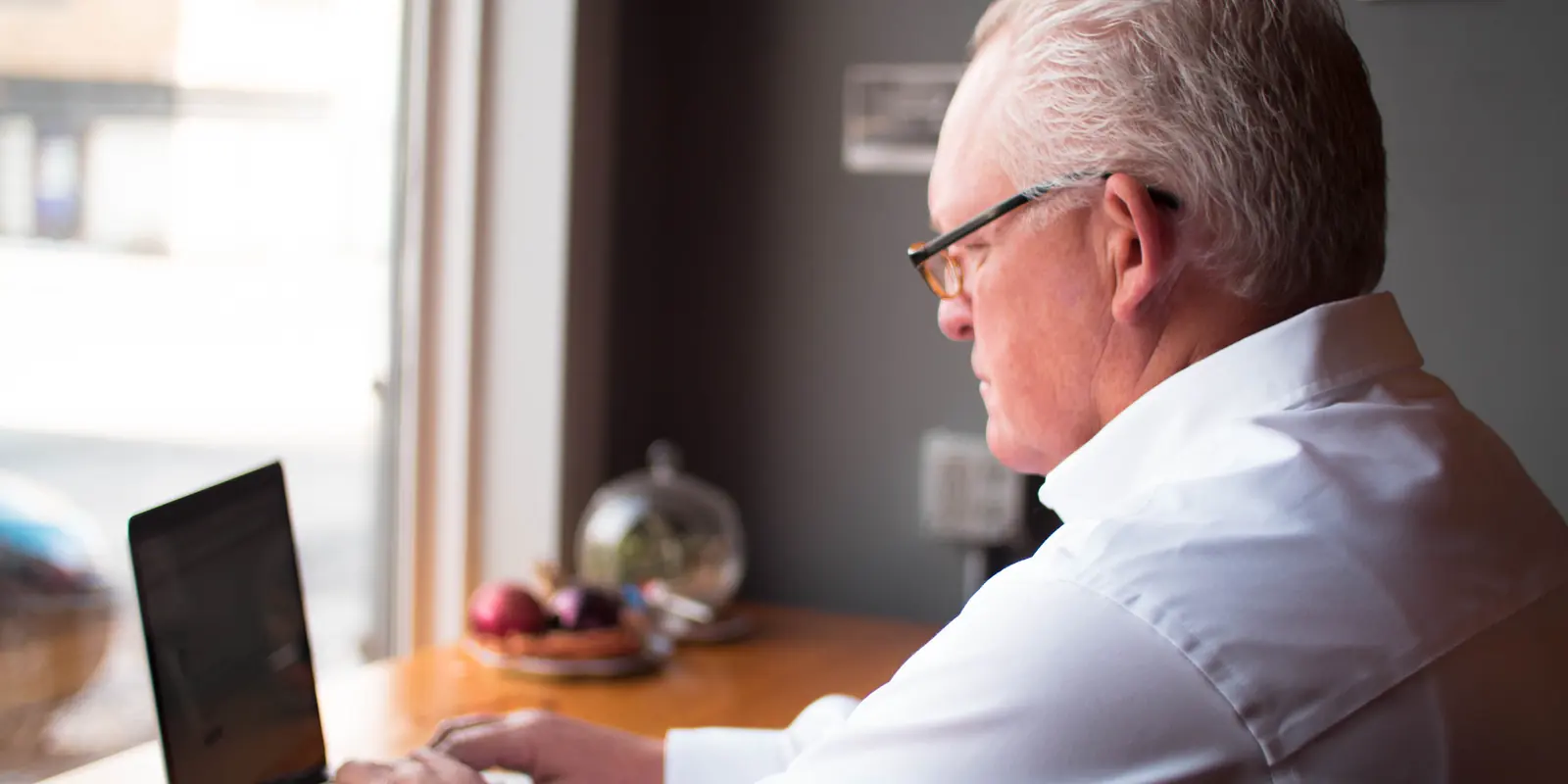 An older man applies online at a laptop by a bright window