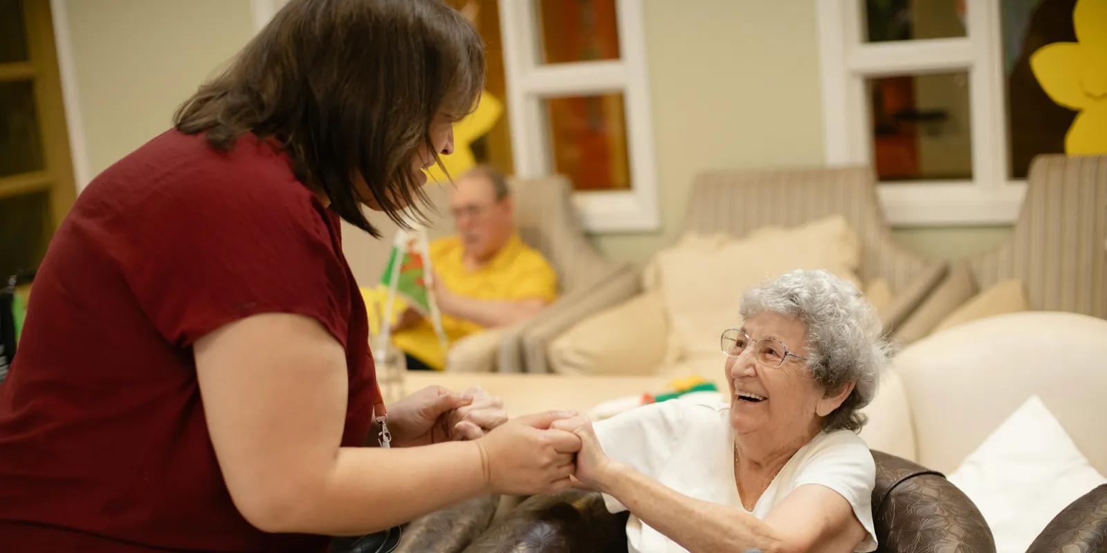 A care worker holds both hands of a smiling older woman seated on a sofa in a care home lounge, with another resident in the background.