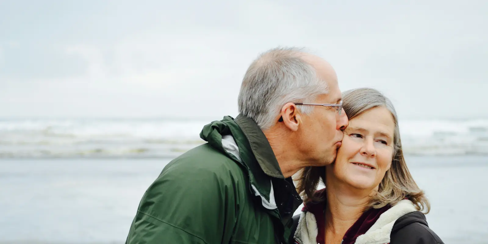 An older couple on an overcast beach, one kissing the other on the cheek