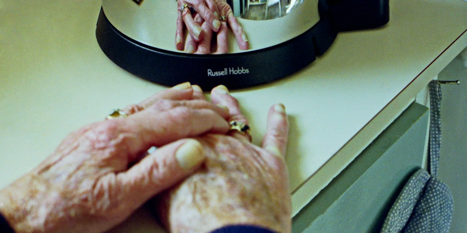 An older woman's hands with wedding rings resting folded on a kitchen counter