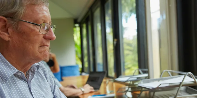 An older man in glasses working at a laptop at a desk by a row of windows