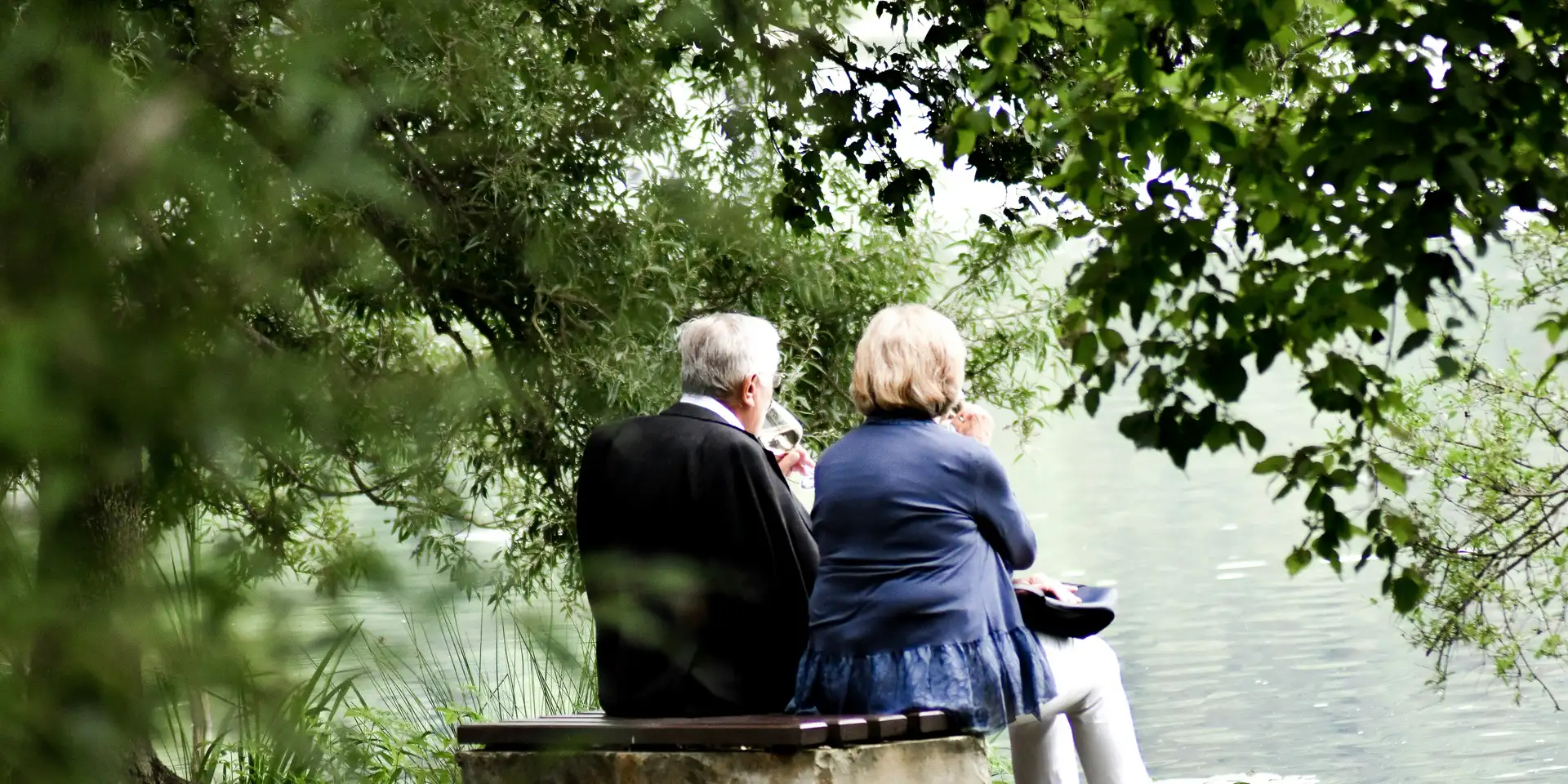 An older couple sitting together on a stone bench beneath trees at the edge of a lake